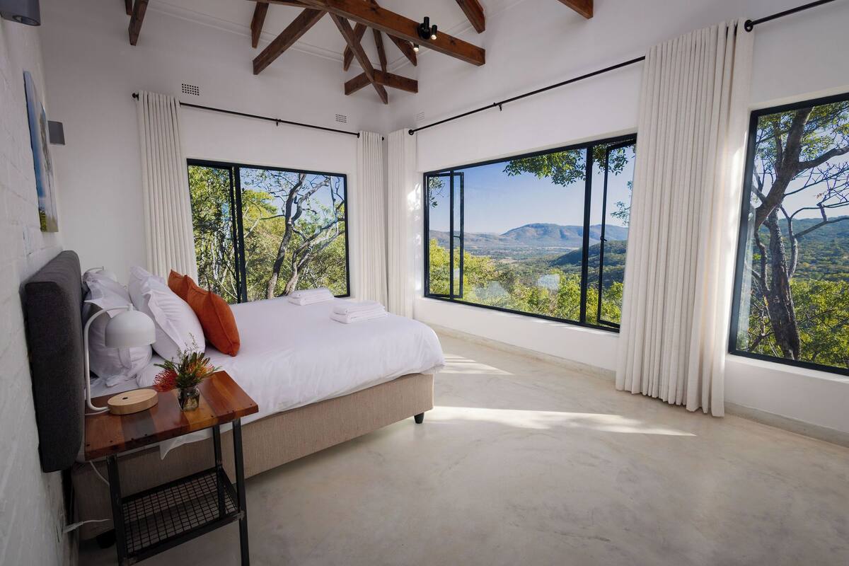 Ein Schlafzimmer mit einem großen Bett, weißen Vorhängen und Blick auf eine Landschaft durch große Fenster.