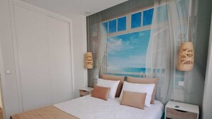 Un dormitorio con una cama grande, vista al mar a través de una ventana y una mesita de noche con lámpara.