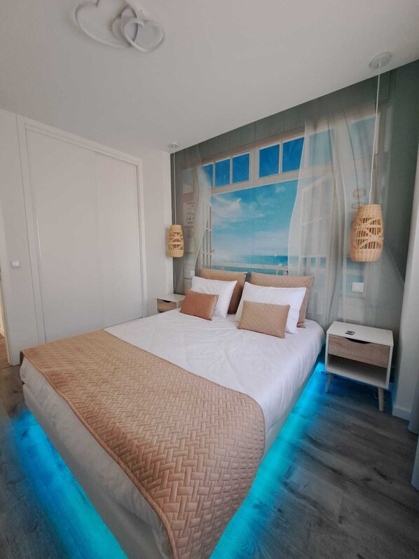 A bedroom with a large bed, a view of the sea through a window, and a nightstand with a lamp.