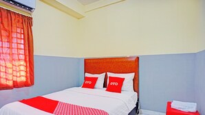 Een hotelkamer met een bed, een rood-wit gebed eiland, twee rode kussens met de merknaam 'OYO', een rode kruk met een handdoek en een raam met oranje gordijnen.