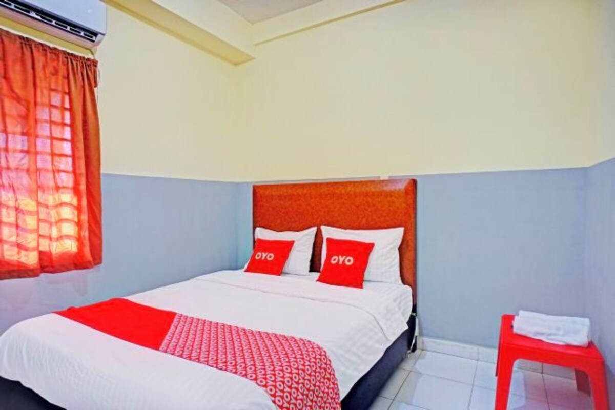 Habitación de hotel con una cama, una colcha roja y blanca, dos almohadas rojas, un taburete rojo y una ventana con cortinas naranjas.