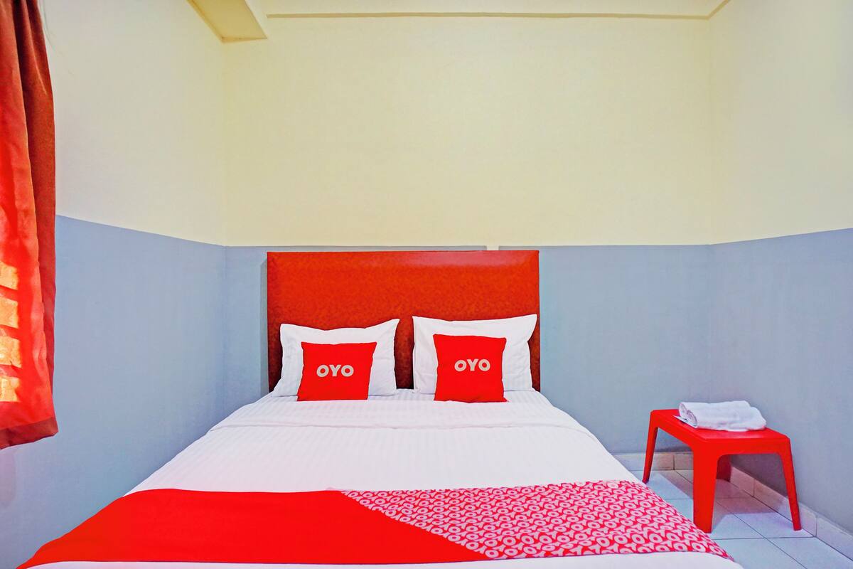 Ein Hotelzimmer mit einem Bett, zwei roten Kissen, einer roten und weißen Tagesdecke, einem roten Hocker und einem weißen Handtuch auf dem Hocker.