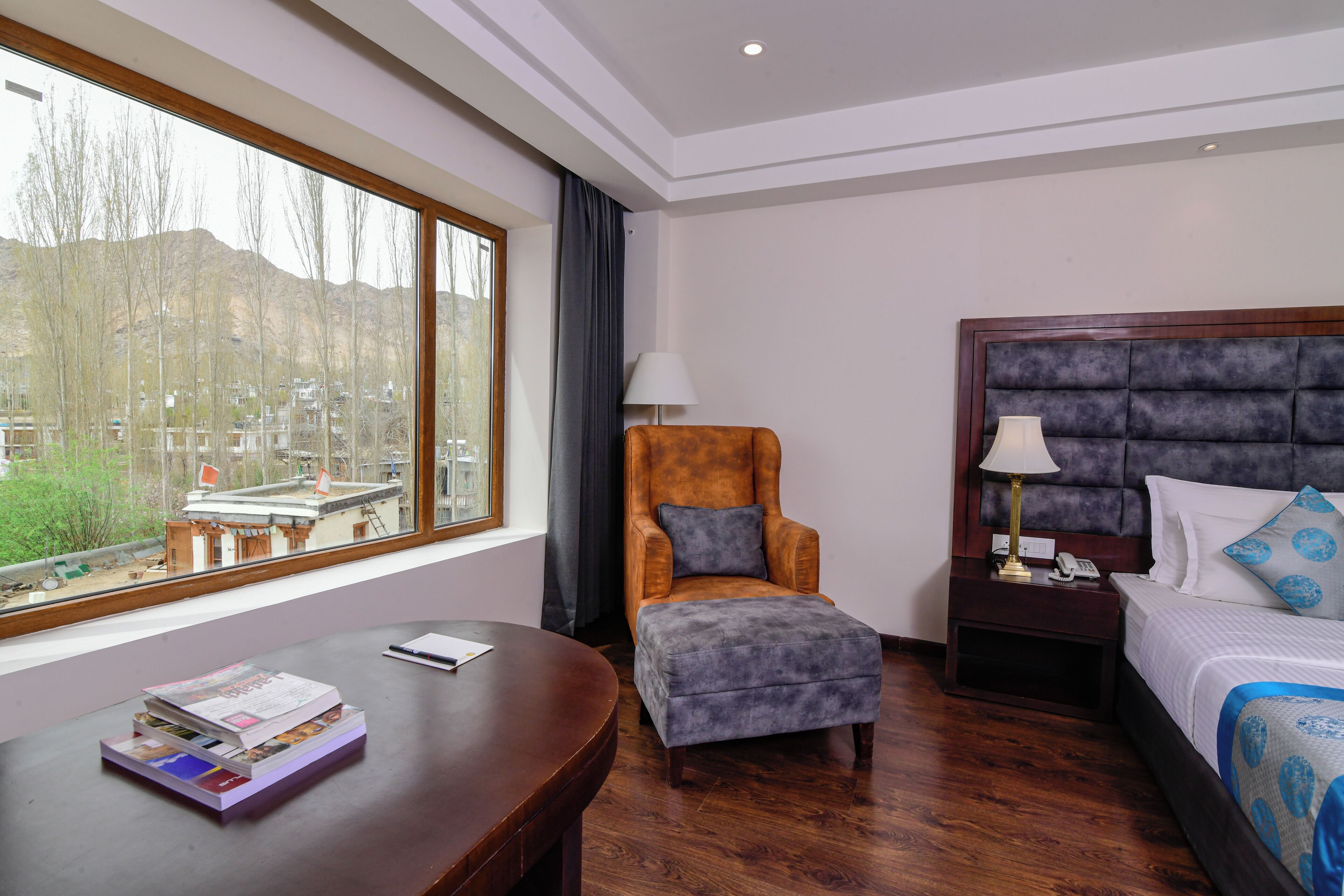 A hotel room with a large window, a bed with a headboard, a chair, a nightstand, and a wooden desk with a lamp.