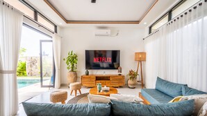 A modern living room with a large flat-screen TV, a comfortable sofa, a coffee table, and a view of a swimming pool.