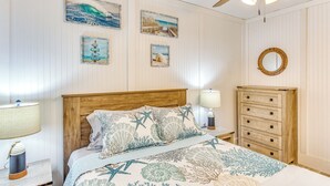 A bedroom with a wooden headboard, a bed with a blue and white patterned bedspread, a nightstand with a lamp, and a dresser with multiple drawers. - Steps to Sand: Remodeled North Myrtle Beach Condo! (North Myrtle Beach)
