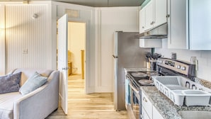 A compact kitchen with a stove, oven, and sink, adjacent to a living area with a sofa and a door leading to another room. - Steps to Sand: Remodeled North Myrtle Beach Condo! (North Myrtle Beach)