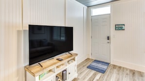 A living room with a flat-screen TV on a wooden stand, a door with a sign, and a blue rug on the floor. - Steps to Sand: Remodeled North Myrtle Beach Condo! (North Myrtle Beach)