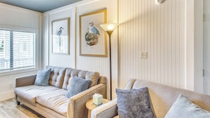 A cozy living room with a beige sofa, a small side table, and a floor lamp. There are framed pictures on the wall and a window with blinds. - Steps to Sand: Remodeled North Myrtle Beach Condo! (North Myrtle Beach)