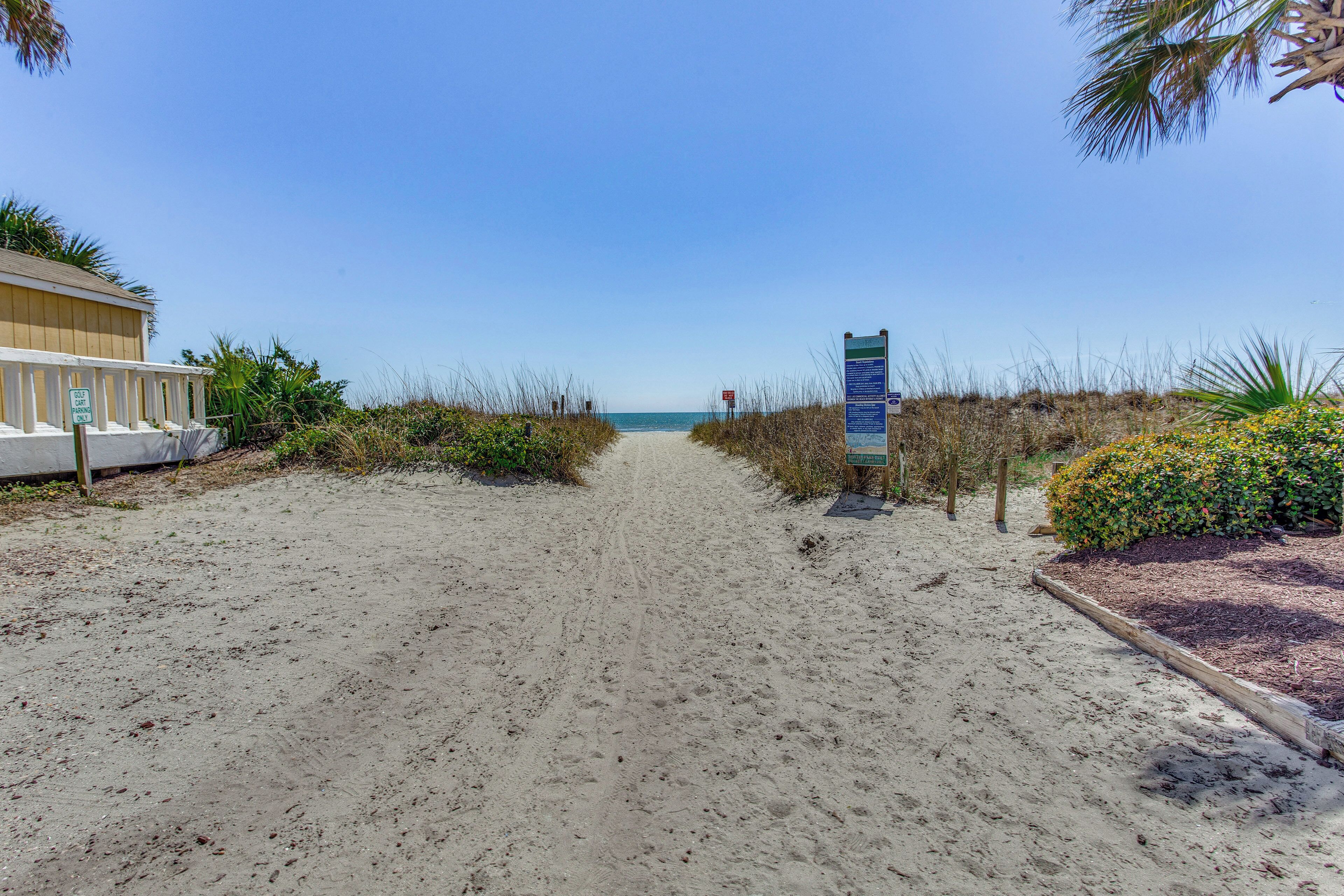 A sandy beach path leading to the ocean, with a signpost and a building on the left.