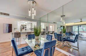 A modern dining area with a glass table and blue upholstered chairs, a large mirror, and a view of the adjacent kitchen and living space. - 8 Mi to Honeymoon Island: Golf Getaway w/ Balcony! (Palm Harbor)