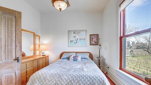A bedroom with a bed, a wooden dresser, a mirror, and a window with a view of a vineyard.