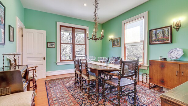 A dining area with a wooden table, chairs, and a chandelier. There are windows with white frames and a view of trees outside.