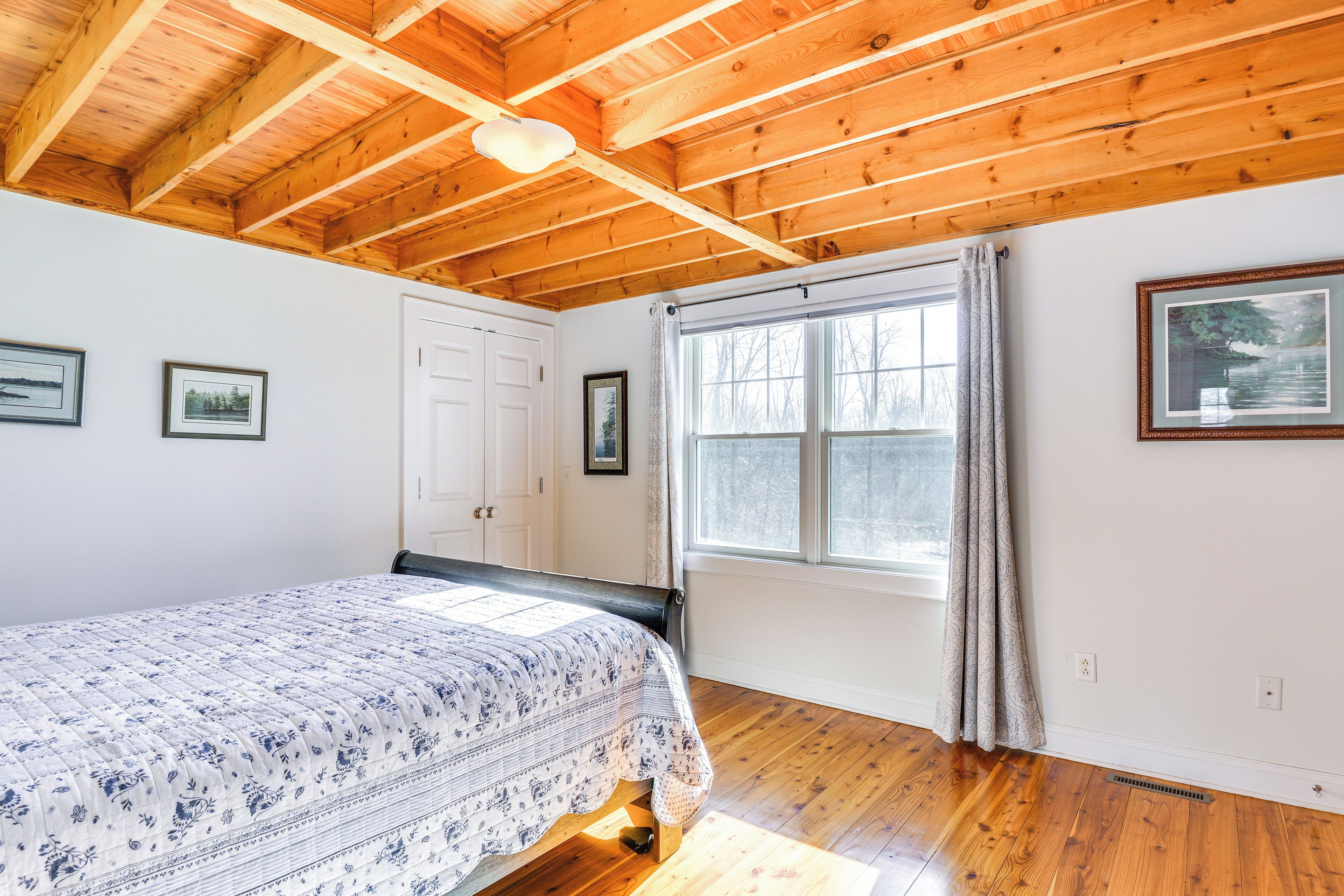 A bedroom with a wooden ceiling, a bed with a patterned bedspread, a window with curtains, and framed pictures on the wall.
