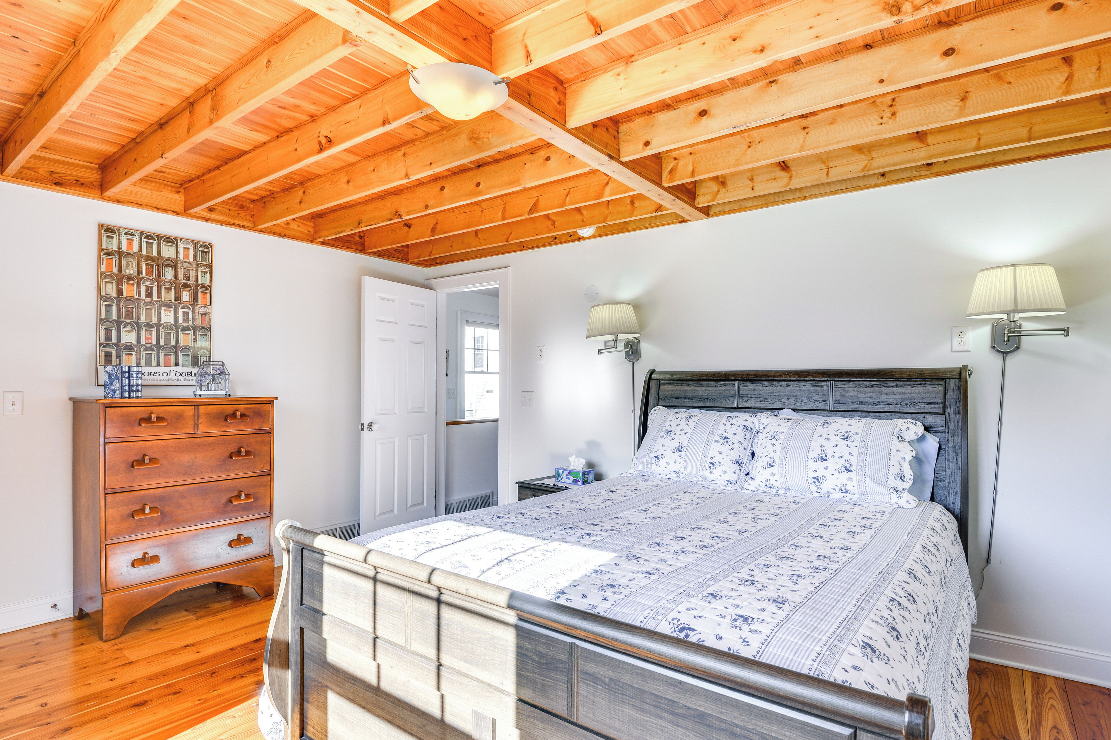 A bedroom with a wooden ceiling, a bed with a floral bedspread, a wooden dresser, and a wall-mounted lamp.