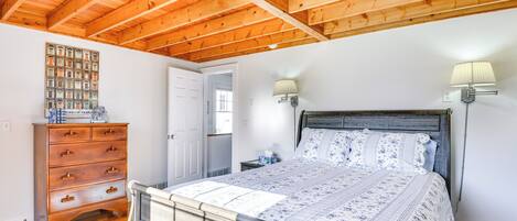 A bedroom with a wooden ceiling, a bed with a floral bedspread, a wooden dresser, and a wall-mounted lamp.