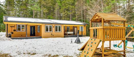 A cabin with a wooden deck and a play structure in a snowy forest.