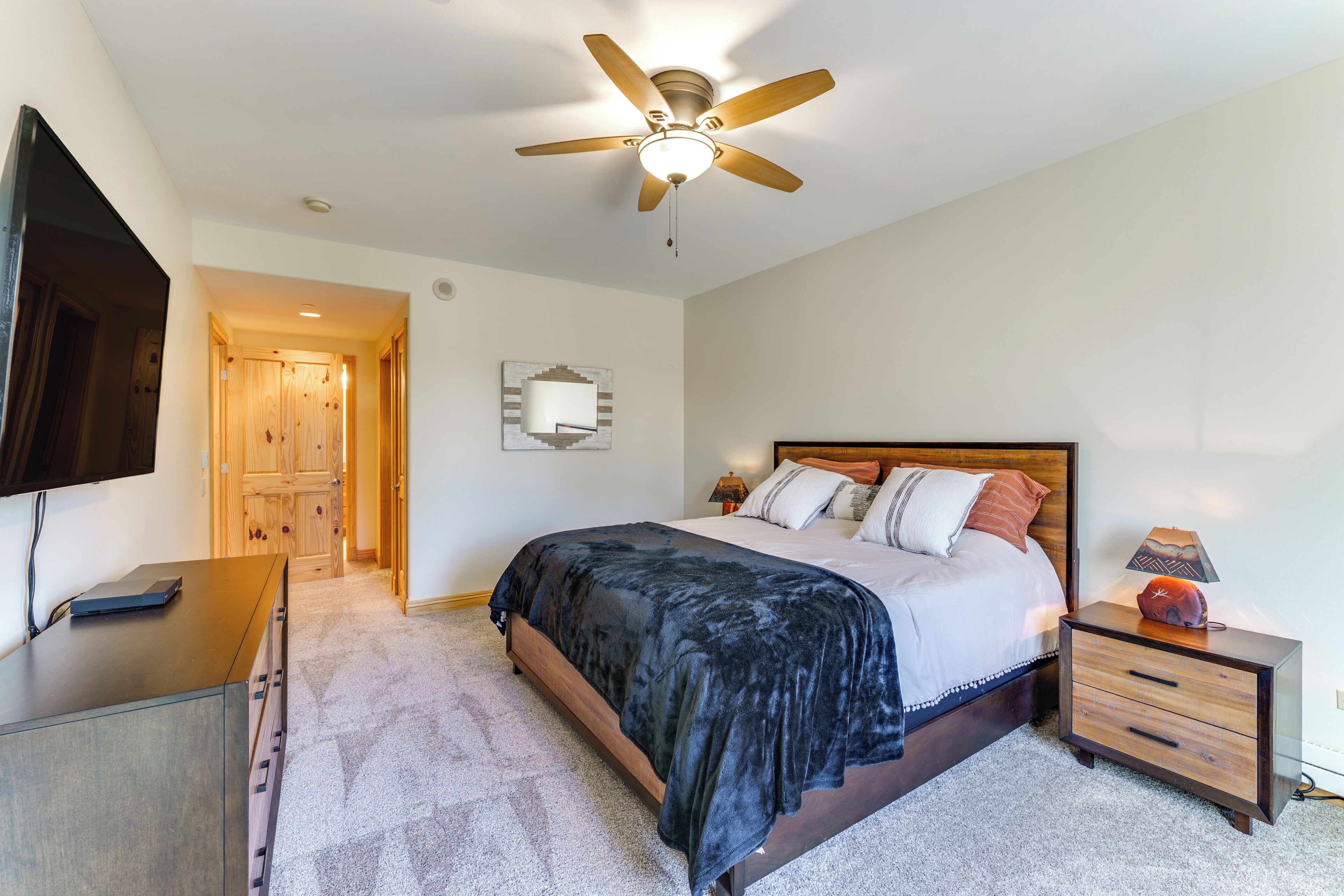 Une chambre à coucher comprenant un lit, une table de chevet, une commode, un ventilateur de plafond et un téléviseur fixé au mur.