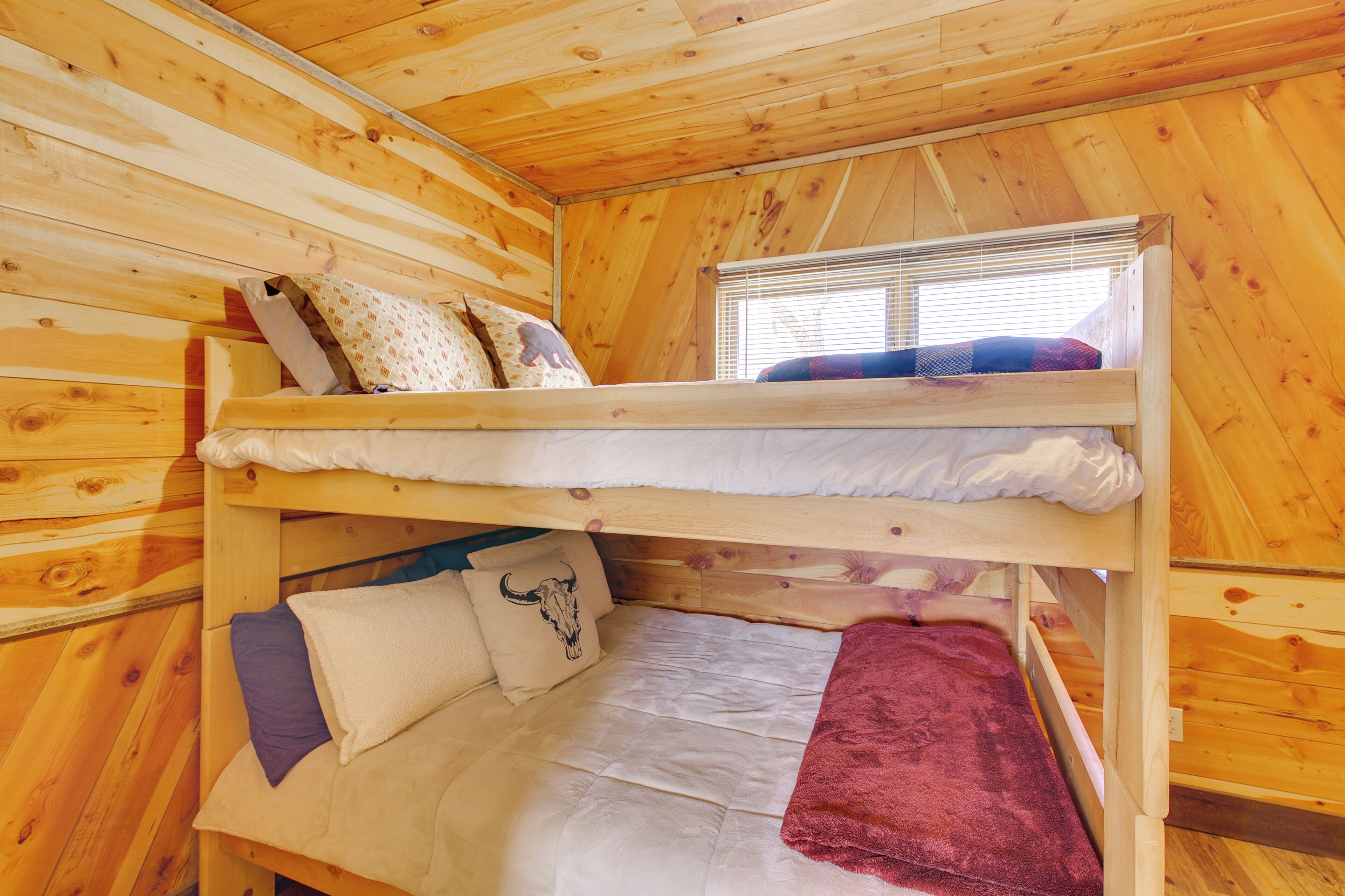 A bunk bed with a window above, wooden walls, and a red blanket on the lower bunk.
