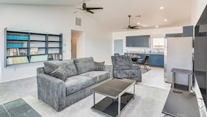 A modern living room with a grey sofa, a coffee table, and a bookshelf. There is a kitchen area in the background with cabinets and a refrigerator.