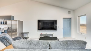 A modern living room with a grey sofa, a flat-screen TV, a dining table, and a kitchen area in the background.