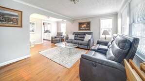A spacious living room with a leather sofa, a glass coffee table, and a rug. There is a painting on the wall and a window with blinds.