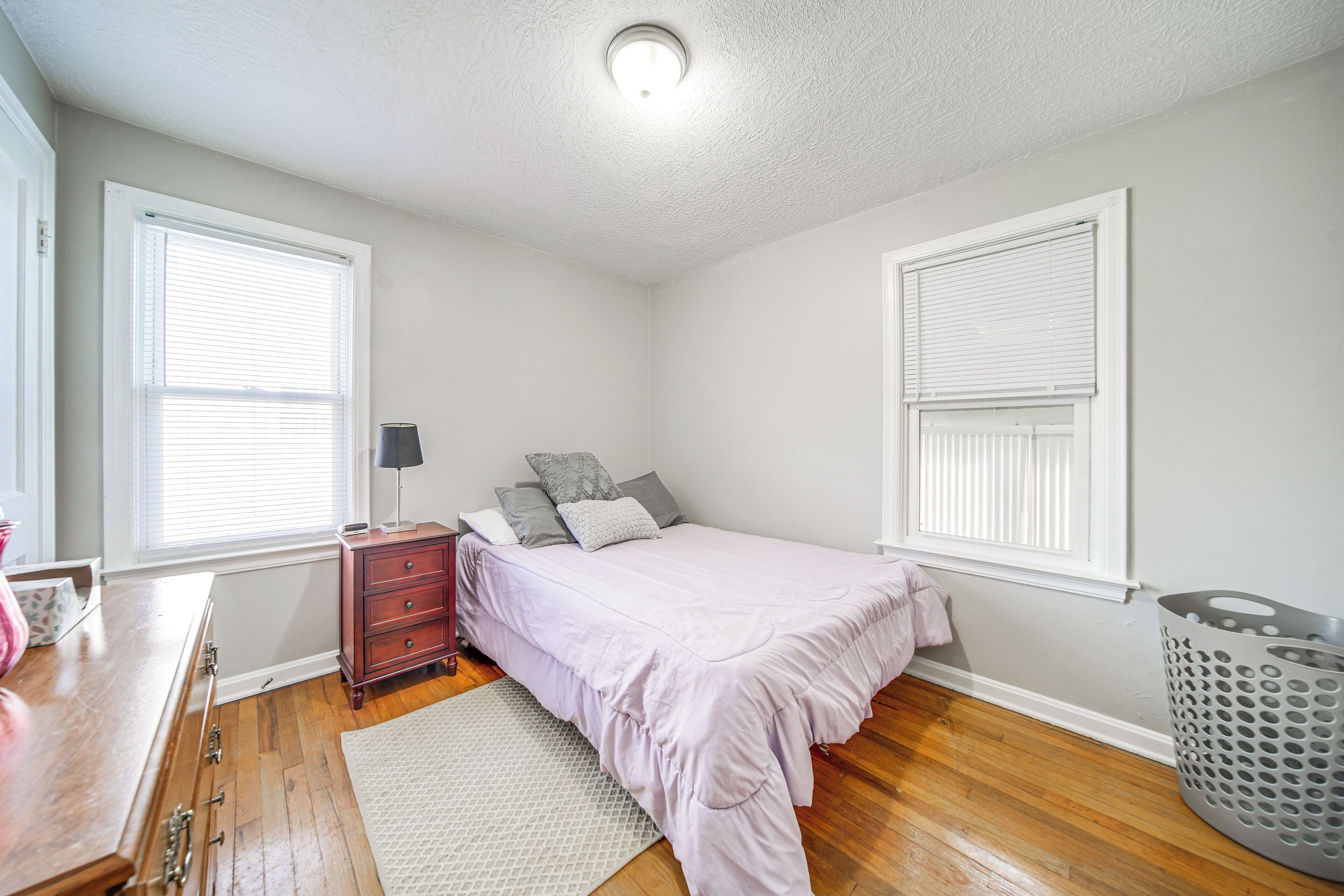 A bedroom with a bed, dresser, nightstand, lamp, and a laundry basket.