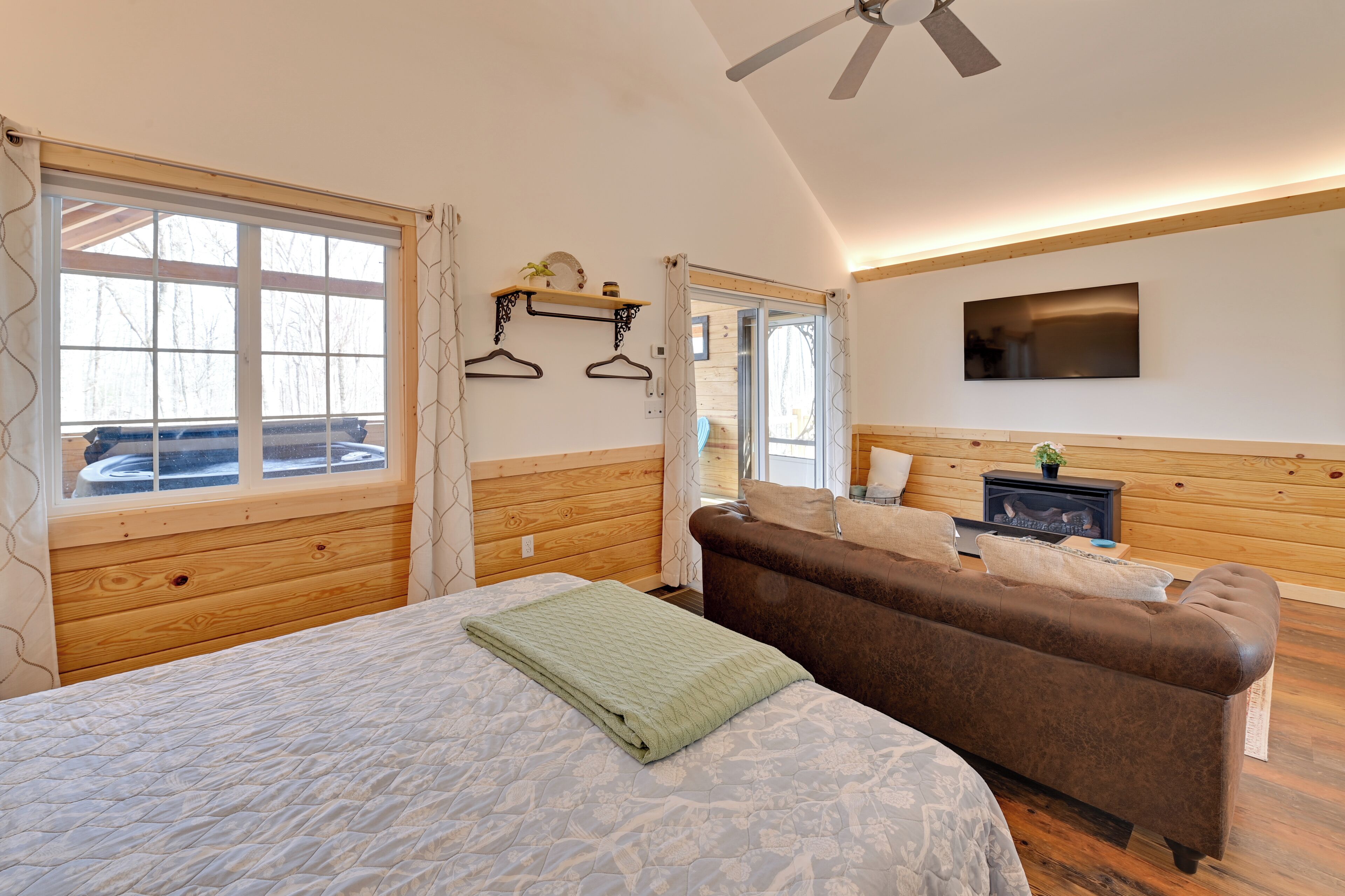 A cozy room with a bed, a brown sofa, a TV, and a window with curtains.