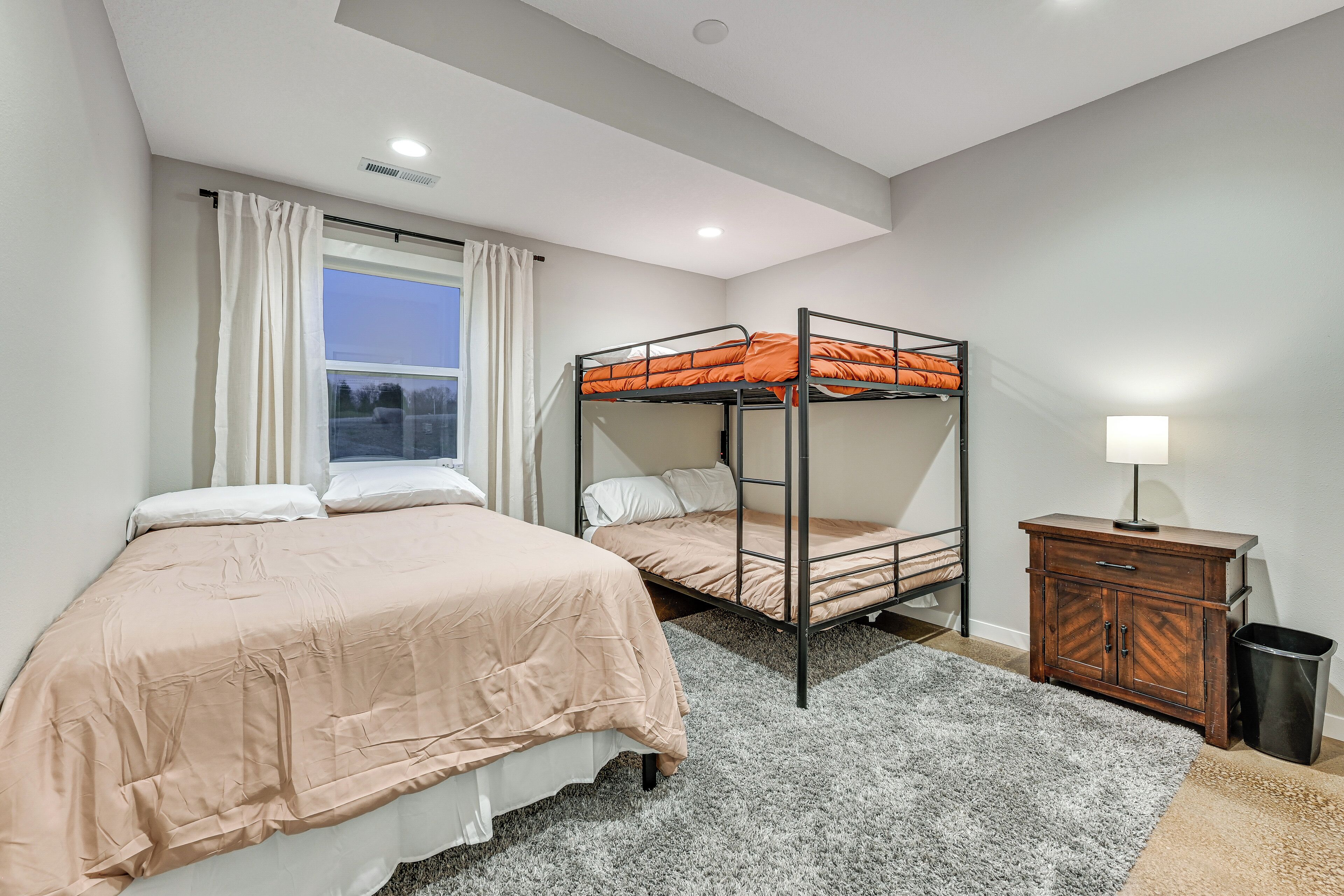 Un dormitorio con una cama litera, una cama individual, un aparador de madera, una lámpara y una ventana con cortinas.