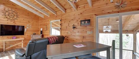 Een houten cabin met een eettafel, bankjes, een bank, een televisie en een groot raam.