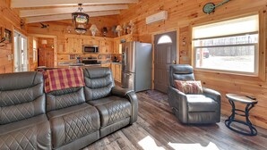 A cozy cabin interior with a leather sofa, a recliner, a dining table, a kitchen area, and a large window. - Plum Crazy: Frenchburg Cabin on 1 Acre (Frenchburg)