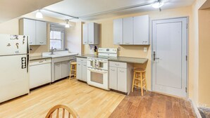 A compact kitchen with white cabinets, a stove, and a sink. There are stools and a wooden chair. - Walkable Studio in the Heart of Pittsburgh! (Pittsburgh)