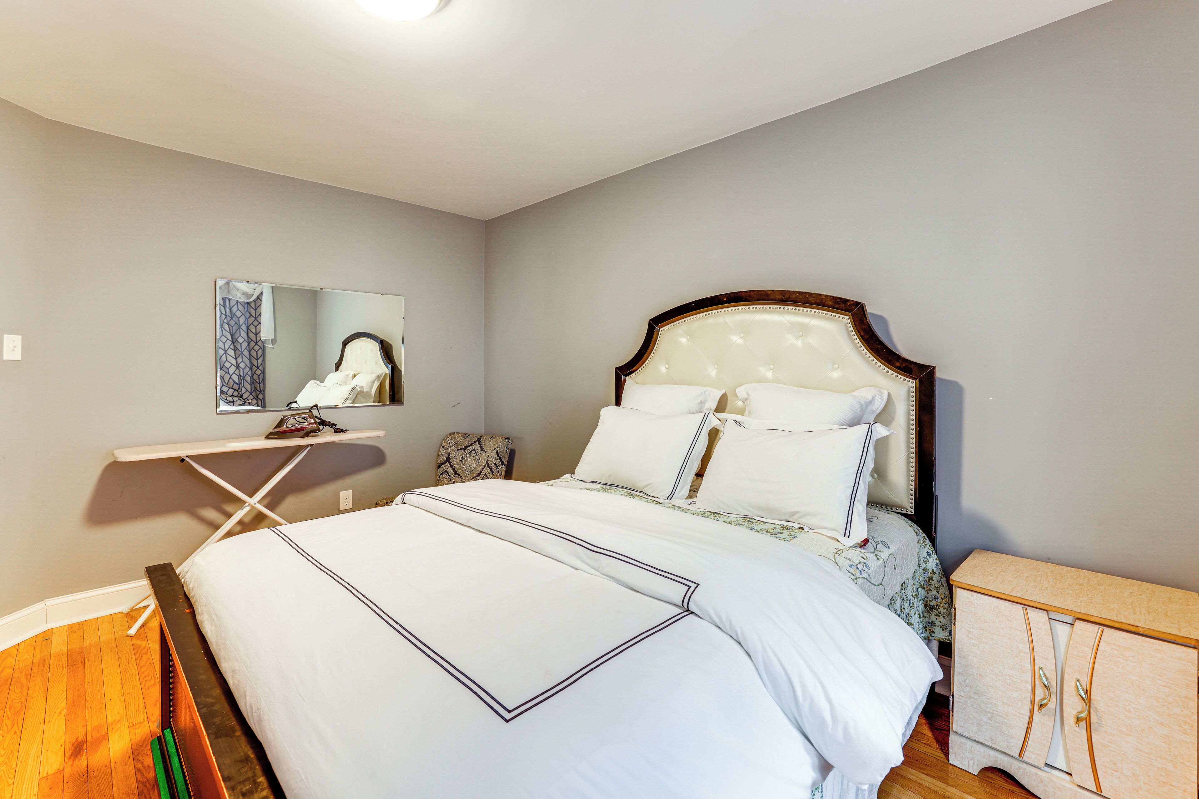 A neatly made bed with white linens and a wooden headboard, a small bedside table with a lamp, and a mirror on the wall.