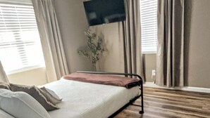 Ein Schlafzimmer mit einem Bett, einem Deckenventilator, einem an der Wand montierten Fernseher und einem Fenster mit Vorhängen.