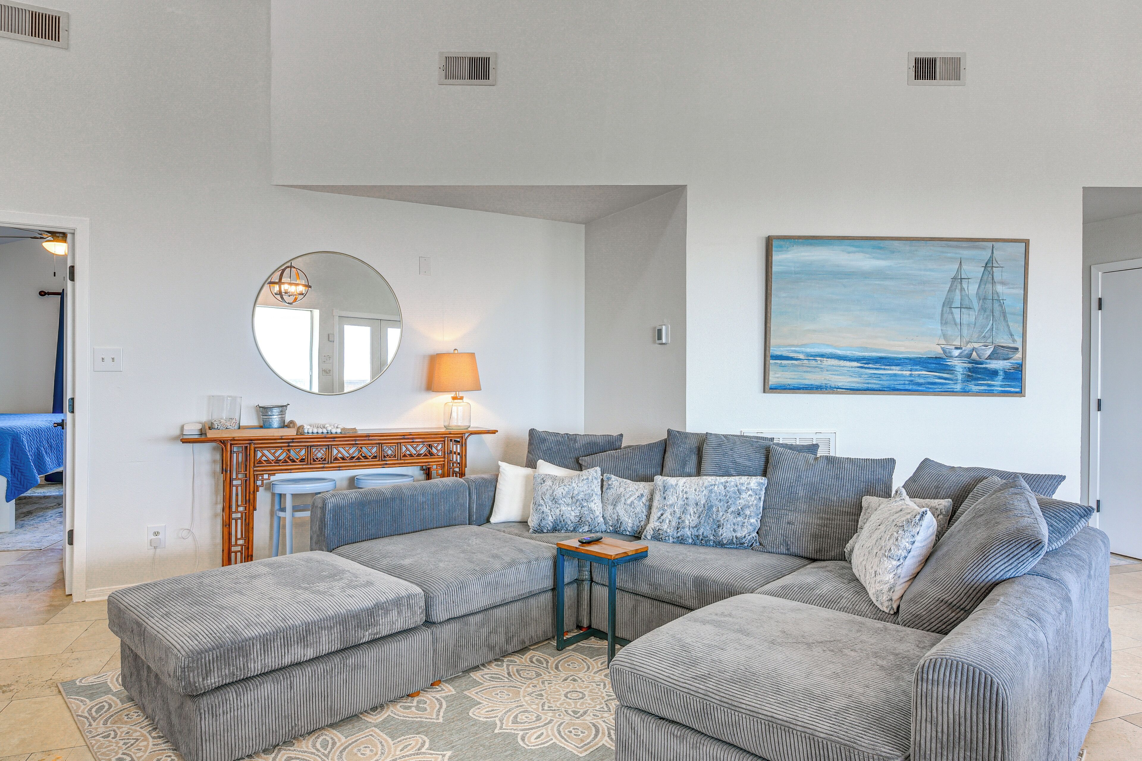 A living room with a sectional sofa, a round mirror, a fireplace, and a painting of a sailboat.
