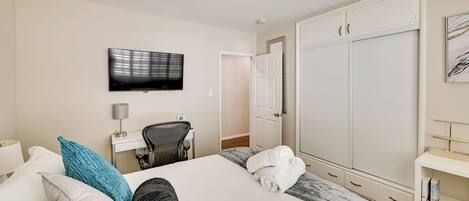 A hotel room with a bed, a TV mounted on the wall, a wardrobe, and a desk with a chair.