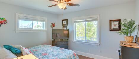 A bedroom with a bed, dresser, ceiling fan, windows, and a plant.