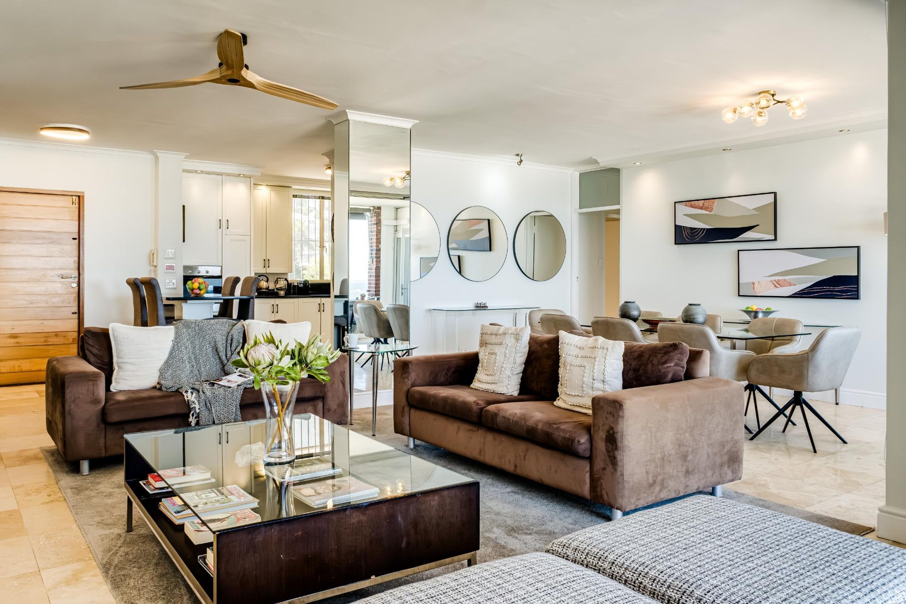 Un salon moderne avec une table basse en verre, des canapés marron et un ventilateur de plafond.