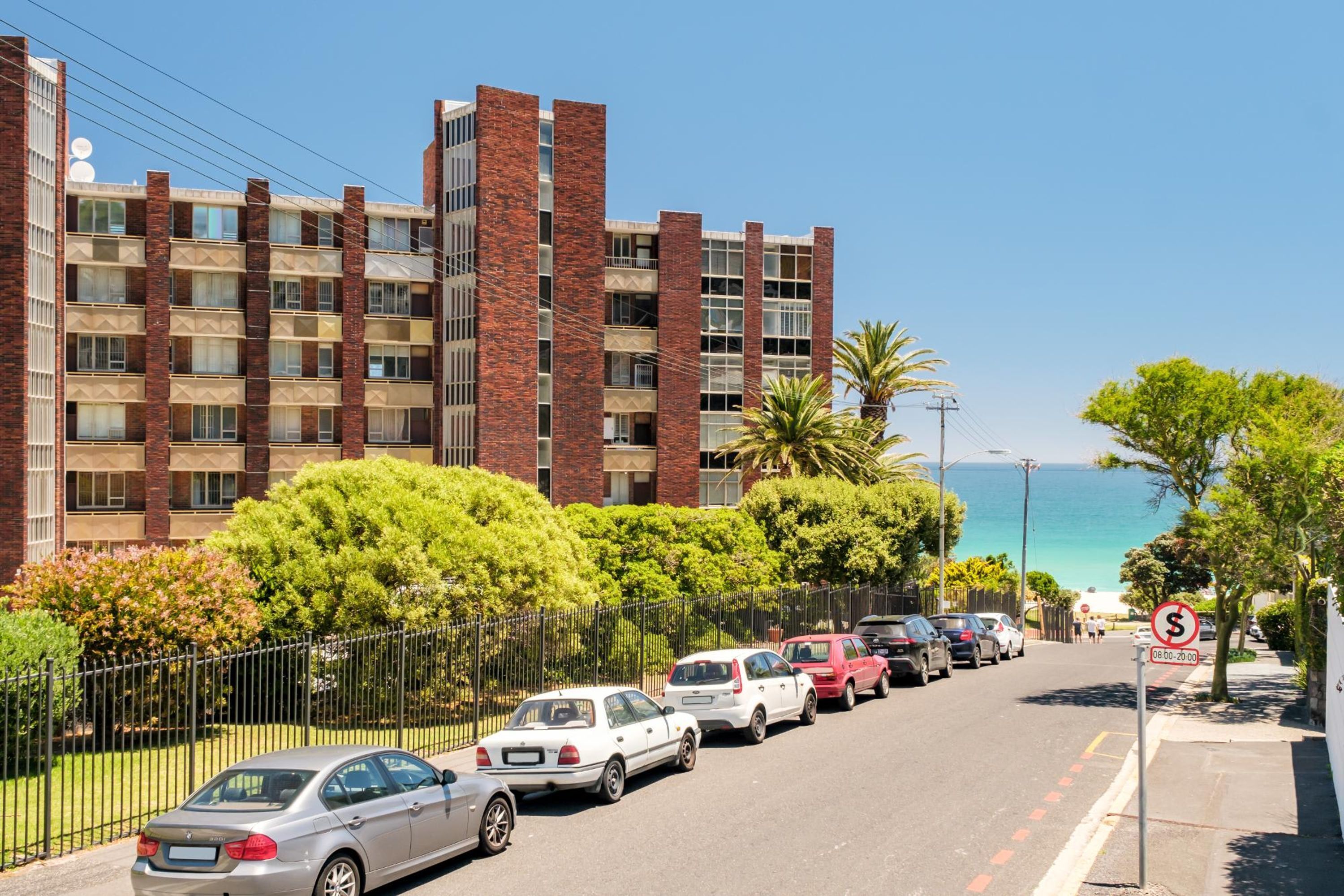 Un edificio de varios pisos con fachada de ladrillo y balcones, situado cerca de la costa, con coches aparcados y un cielo despejado.