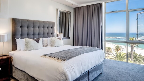 A bedroom with a large bed, a window with a view of the beach, and a grey headboard. - Camps Bay Beachside 2bed w Sea View Cloud 9 (Cape Town)