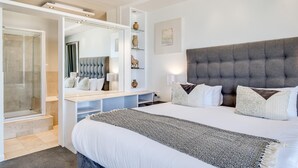 A modern bedroom with a large bed, a grey tufted headboard, a walk-in closet, and a glass shower enclosure. - Camps Bay Beachside 2bed w Sea View Cloud 9 (Cape Town)