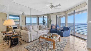 A spacious living room with a large window, a ceiling fan, a sofa, a coffee table, and a blue armchair. - ☀️Ebb Tide 806-Oceanfront with Fabulous Views-Pool+Hot Tub☀️ (New Smyrna Beach)