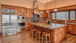 A modern kitchen with wooden cabinets, stainless steel appliances, a large island with stools, and a stone backsplash. - Incredible Panoramic View, Close to Spearfish, Very Open Floor Plan & Paved! (Spearfish)