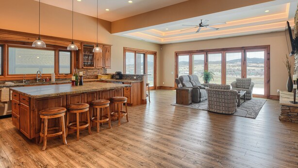 A spacious kitchen with a large island and bar stools, a dining area with a sofa and chairs, and large windows offering a view of the landscape. - Incredible Panoramic View, Close to Spearfish, Very Open Floor Plan & Paved! (Spearfish)