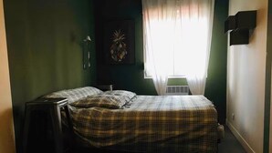 A small, dimly lit room with a single bed, a nightstand, a wall-mounted lamp, and a window with sheer curtains. - Nice house in the center of Arès (Unknown)