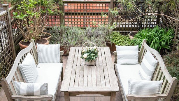 A wooden deck with a table and two cushioned benches, surrounded by potted plants and a wooden fence.
