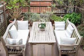 A wooden deck with a table and two cushioned benches, surrounded by potted plants and a wooden fence.