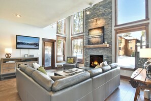 Ein geräumiges Wohnzimmer mit Kamin aus Naturstein, großen Fenstern, einer gemütlichen Couch und einem flachbildfernseher an der Wand.