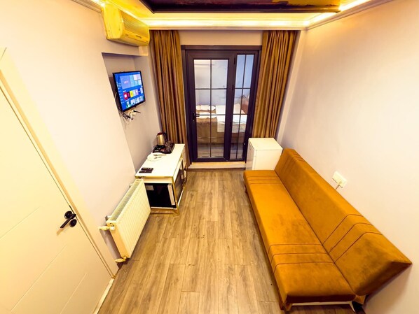 A compact room with a wooden floor, a sofa, a small table with a kettle, a radiator, a TV on the wall, and a sliding glass door leading to a balcony. - Brick House Hotel (Istanbul)
