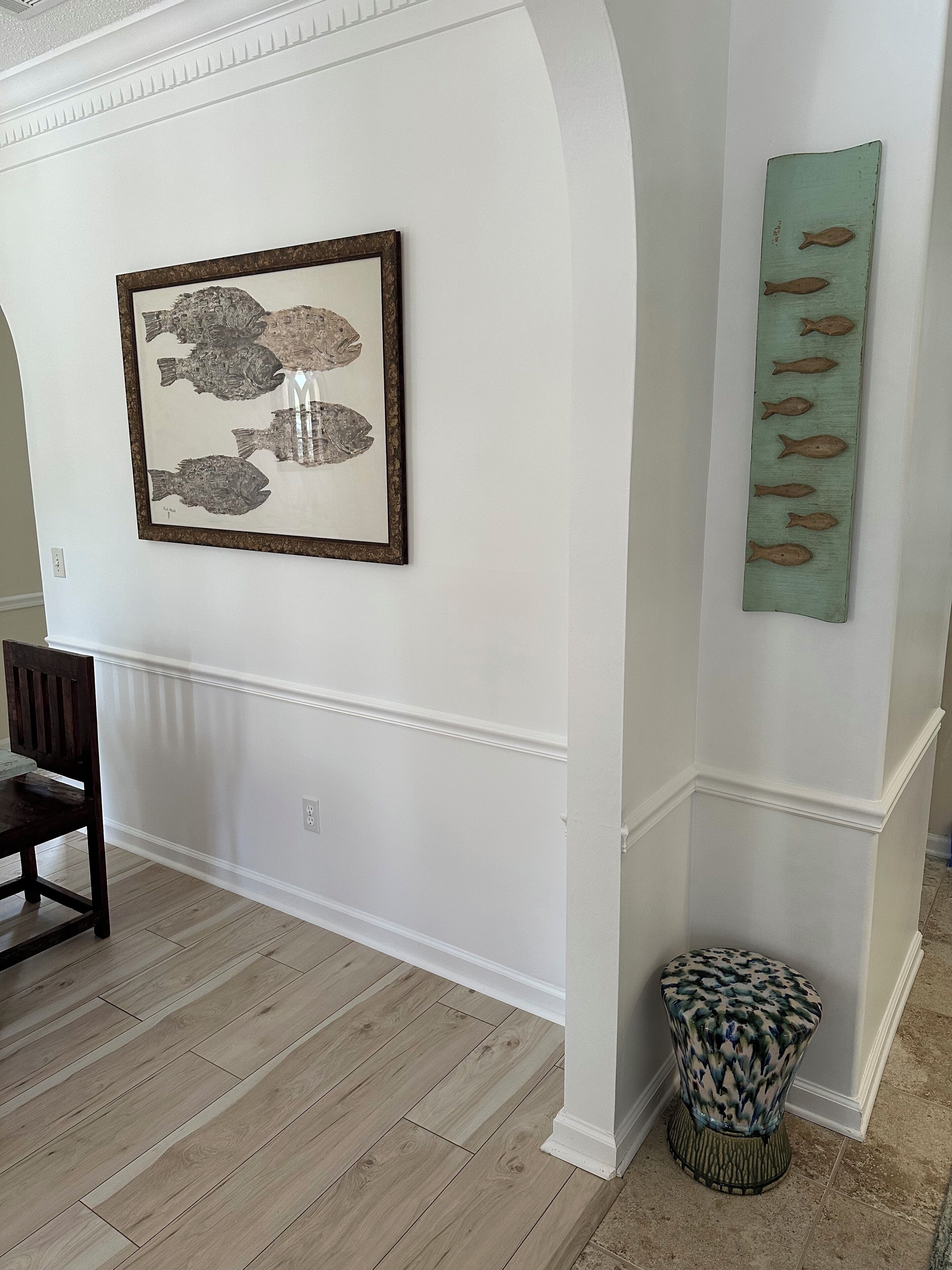 A room with a framed fish artwork, a decorative fish wall piece, a wooden chair, and a patterned stool.