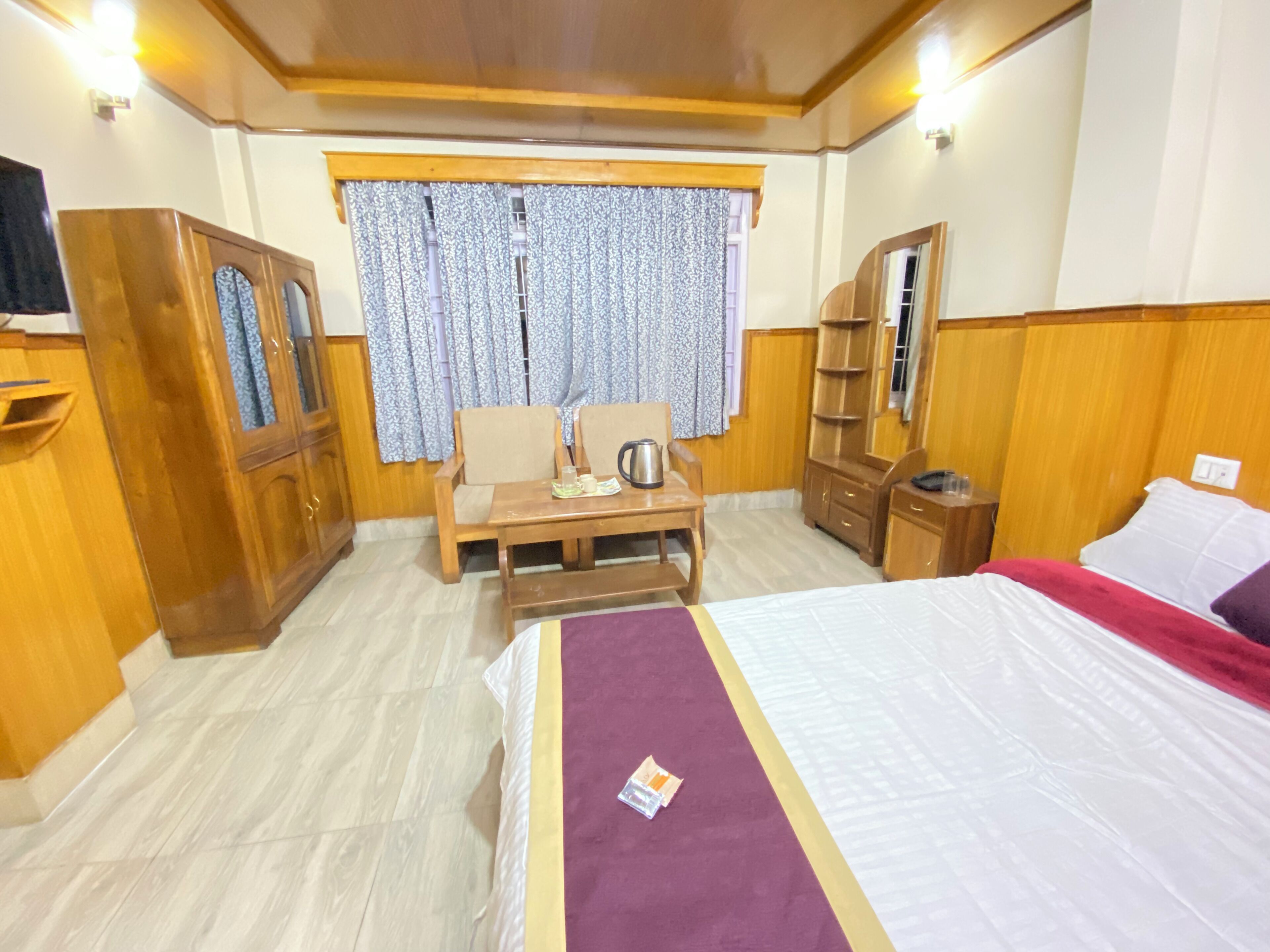 Habitación de hotel con una cama, un escritorio de madera, una silla, una mesita con una tetera, un armario y una ventana con cortinas.